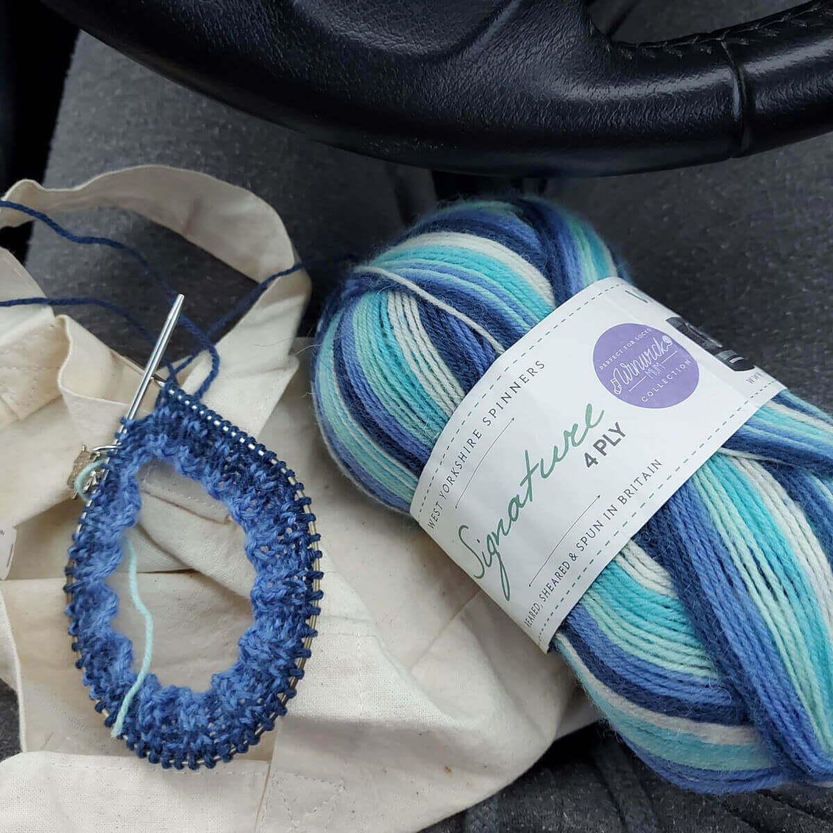 A cream bag is resting on Christine's knee with a short circular needle on the left, with a few rounds of rib knitting on it. The yarn is shades of blue. To the right is the ball of yarn which is striped yarn in shades of blue, pale green, navy, turquoise and cream.