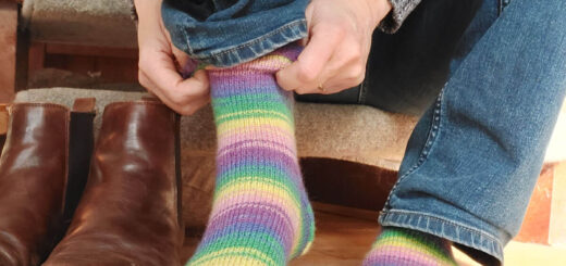 Christine is sitting on the bottom step of the staircase putting on a pair of pink, blue, green, purple and yellow striped socks. There is a pair of brown boots on the wooden floor next to her.