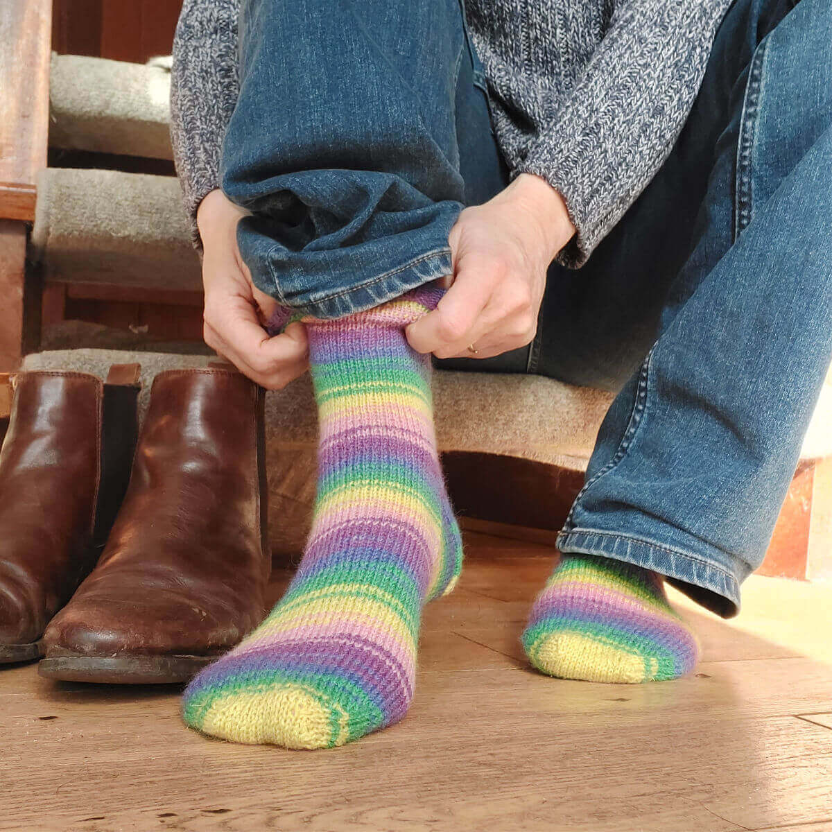 Christine is sitting on the bottom step of the staircase putting on a pair of pink, blue, green, purple and yellow striped socks. There is a pair of brown boots on the wooden floor next to her.