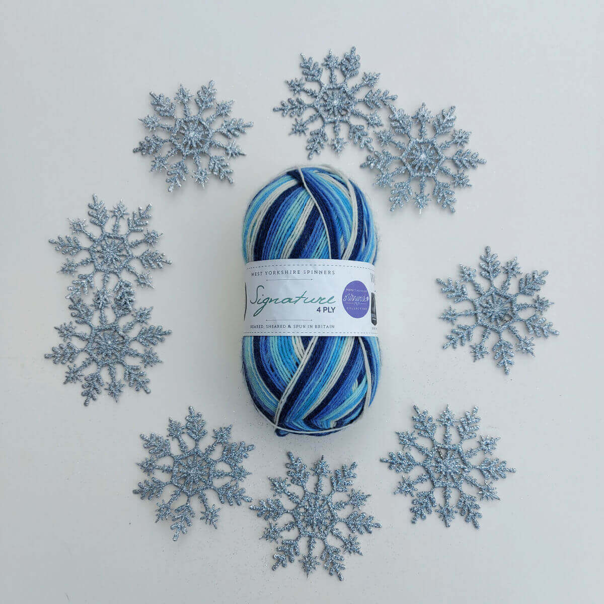 A ball of blue, navy, turquoise and cream striped yarn is on a white background surrounded by silver snowflakes