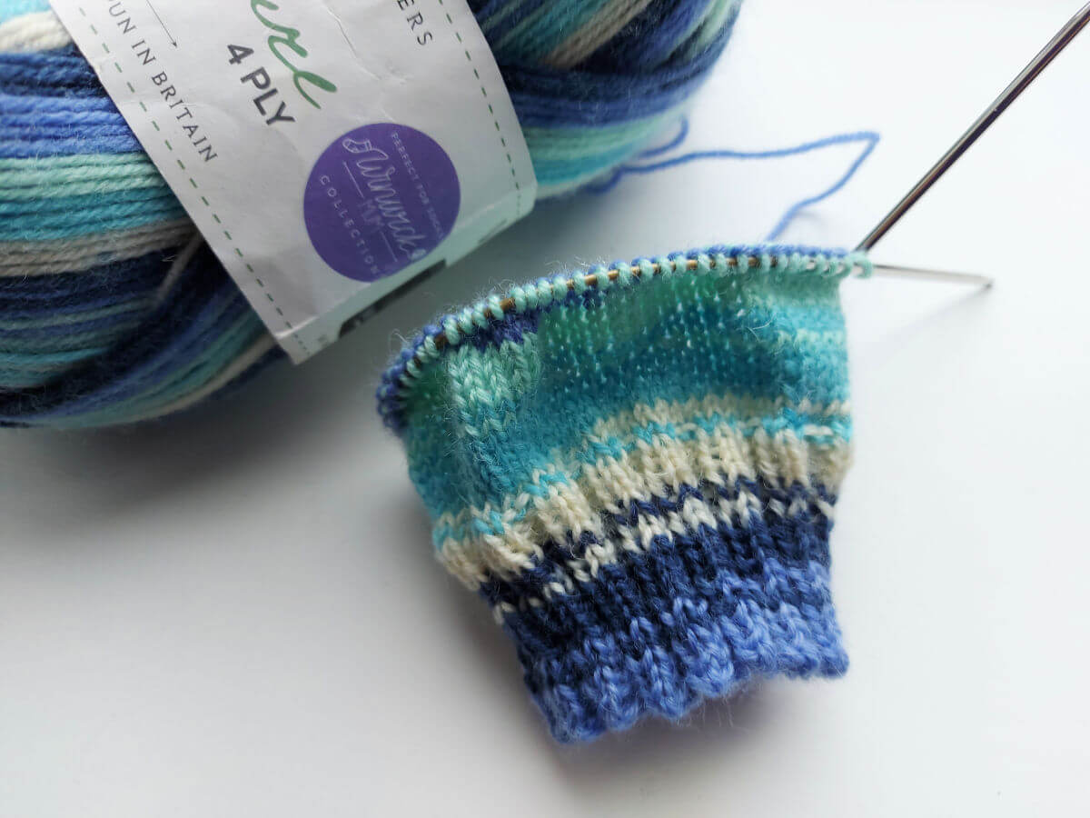 A partly-knitted sock in shades of blue, turquoise and cream on a short circular needle