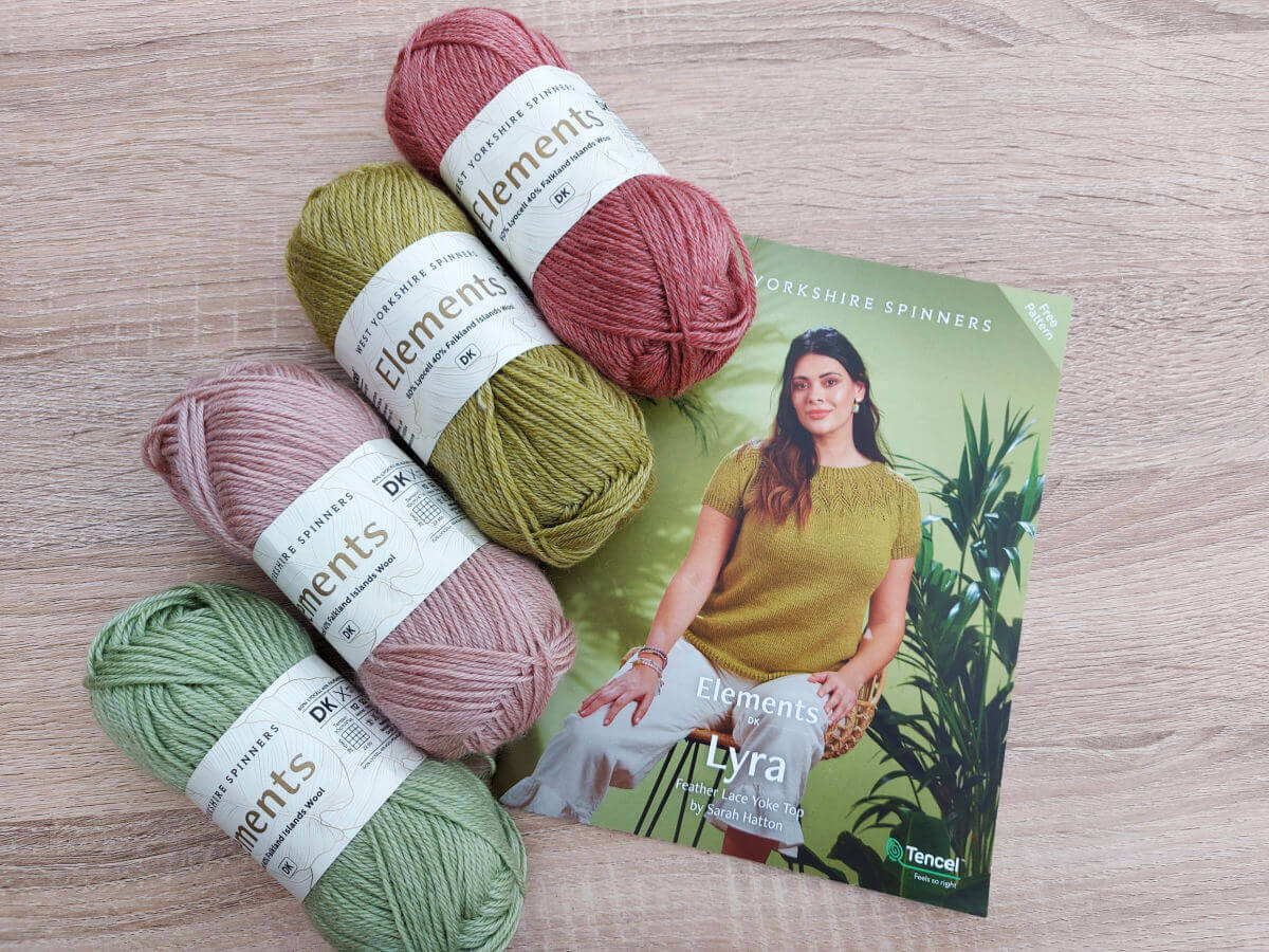 Four balls of yarn - two shades of pink and two shades of green - on a wooden table next to a knitting pattern of a short-sleeved lacy top