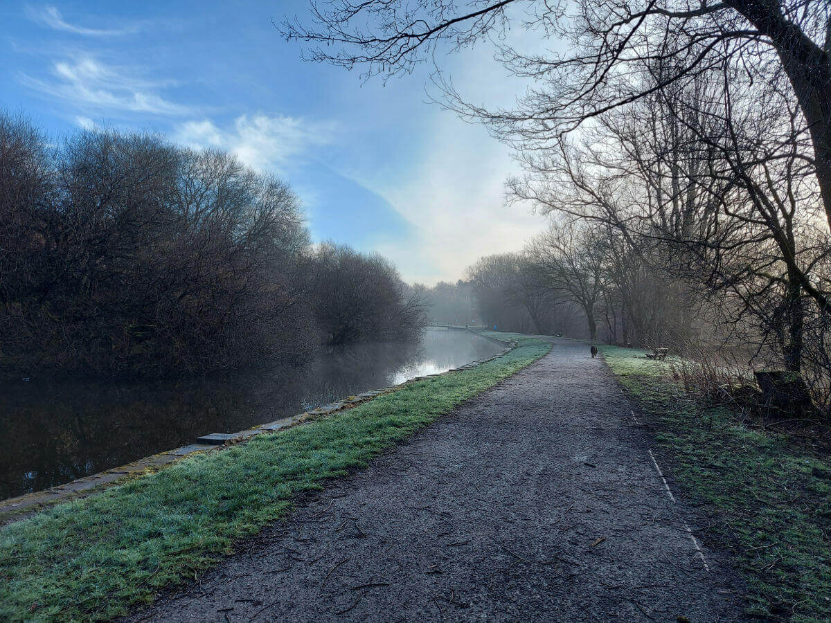 A black dog is in the distance along a canal tow path. The sky is blue with wispy clouds.