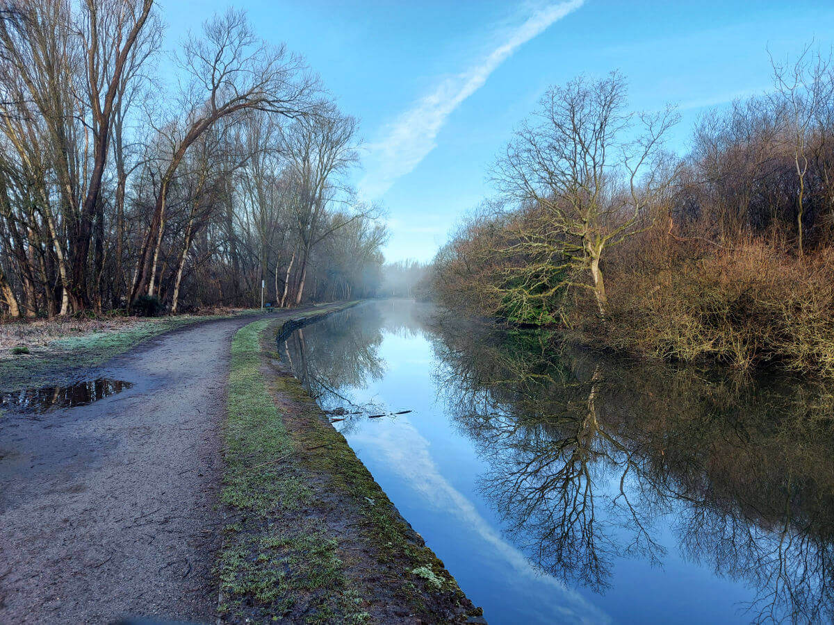 A bright blue sky above a canal. The sky and an aeroplane vapour trail are reflected in the water as well as the trees along the canal bank