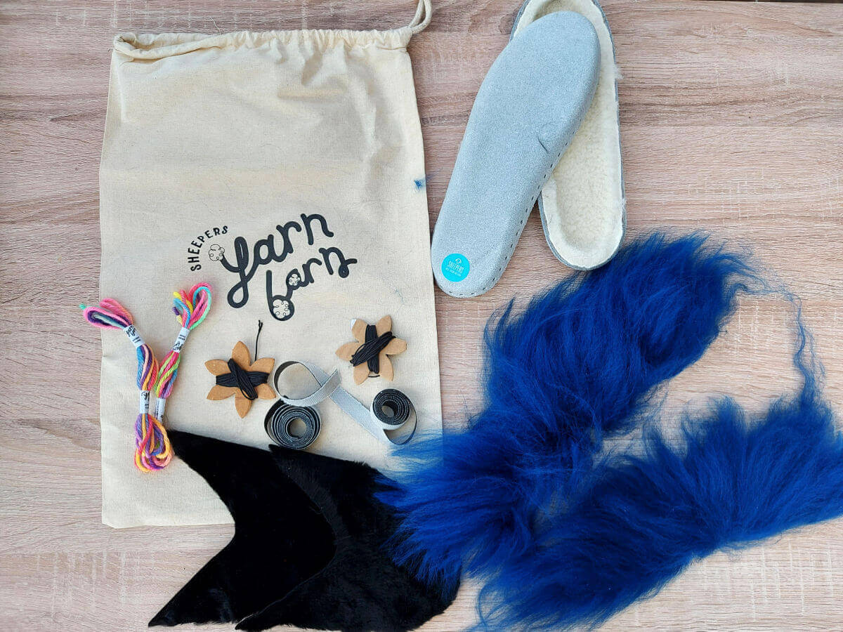 An undyed fabric bag, two small skeins of coloured wool, black slipper front shapes, two pre-made slipper bases and two blue sheepskin strips on a wooden table
