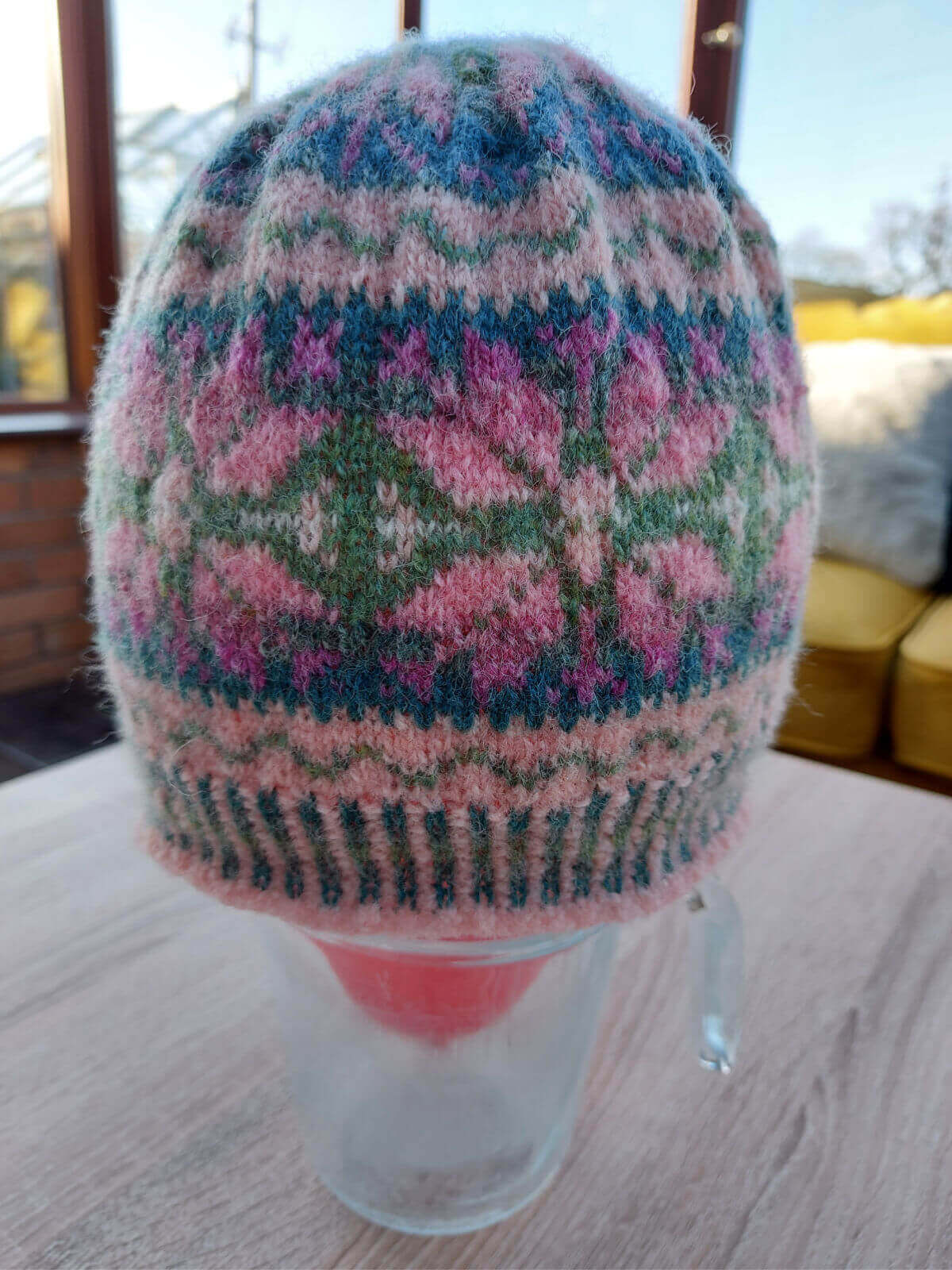 A knitted colourwork hat is drying on a red balloon balance in a glass jug. The jug is standing on a wooden table