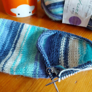 A blue, turquoise and white striped sock lies on a wooden table next to an orange mug with a sheep head on it and a ball of yarn in the same colour