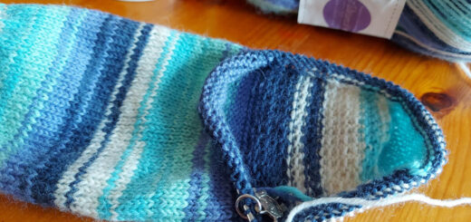 A blue, turquoise and white striped sock lies on a wooden table next to an orange mug with a sheep head on it and a ball of yarn in the same colour