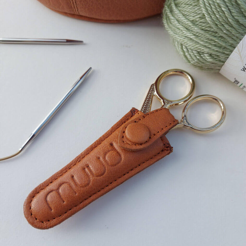A pair of gold scissors in a brown leather Muud Espoo scissors case. There's a ball of green yarn, the edge of a brown leather project bag and a circular knitting needle surrounding the scissors.