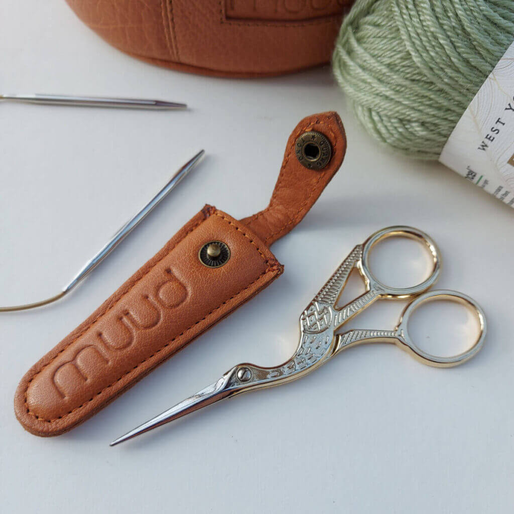 A pair of gold scissors next to a brown leather Muud Espoo scissors case. There's a ball of green yarn, the edge of a brown leather project bag and a circular knitting needle surrounding the scissors.