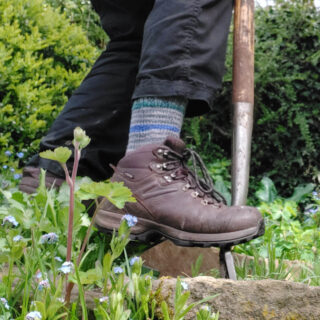 Christine is digging in the garden with a garden fork. She is wearing battered brown boots, black trousers and a pair of grey, green and blue striped socks