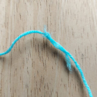 A strand of turquoise blue yarn lies on a pale wooden table. The yarn has broken and been plied together again, leaving fluffy edges at either end of the join.