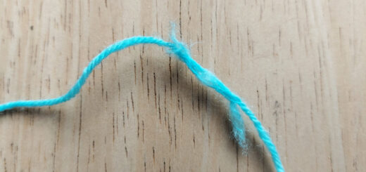 A strand of turquoise blue yarn lies on a pale wooden table. The yarn has broken and been plied together again, leaving fluffy edges at either end of the join.