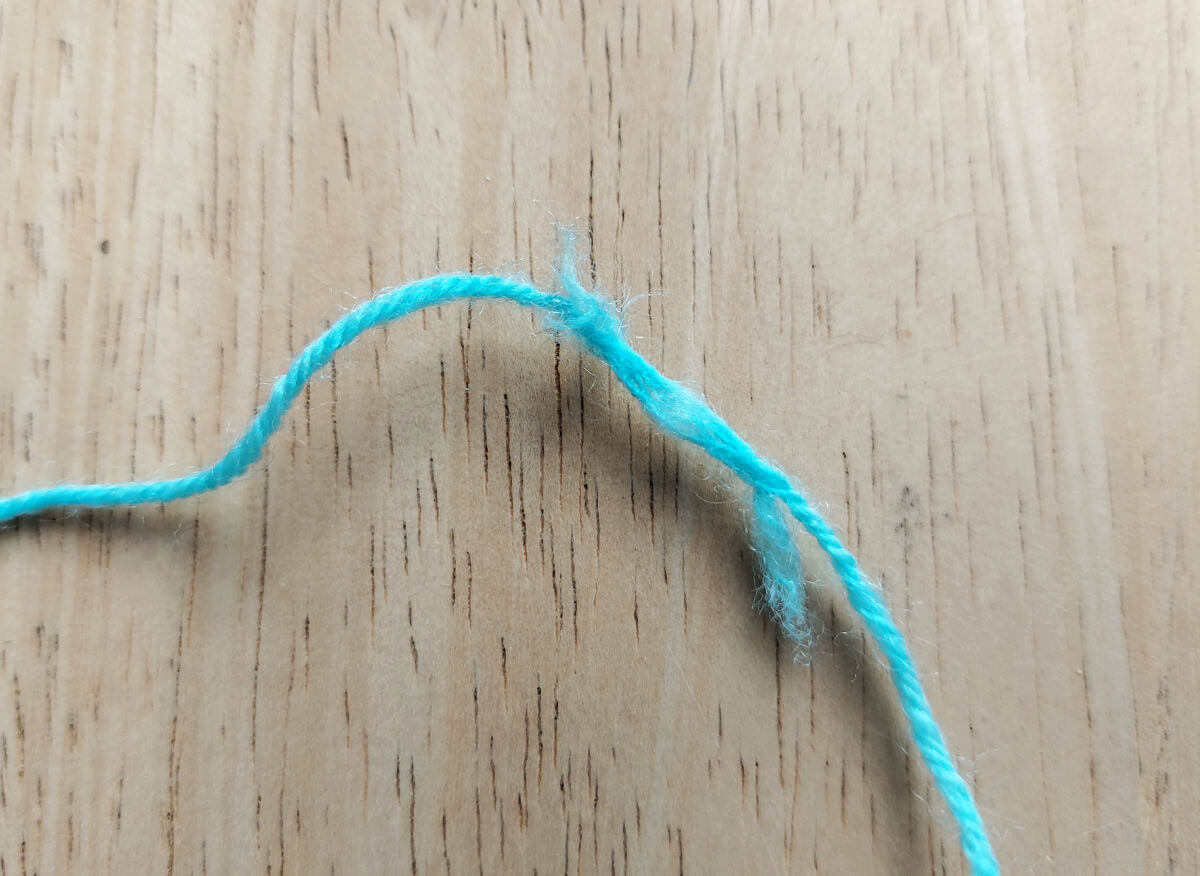 A strand of turquoise blue yarn lies on a pale wooden table. The yarn has broken and been plied together again, leaving fluffy edges at either end of the join.
