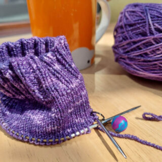 A partly-knitted sock in purple yarn is sitting on a wooden table. In the background is the cake of purple yarn and an orange mug.