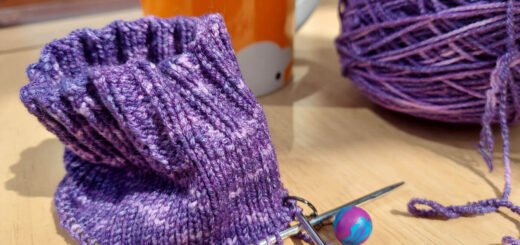 A partly-knitted sock in purple yarn is sitting on a wooden table. In the background is the cake of purple yarn and an orange mug.