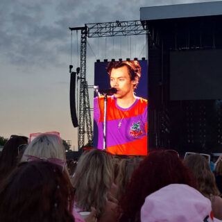 A view across a crowded stadium towards a stage and a giant screen showing a picture of Harry Styles. He is wearing a red and purple t-shirt and singing into a microphone