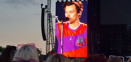 A view across a crowded stadium towards a stage and a giant screen showing a picture of Harry Styles. He is wearing a red and purple t-shirt and singing into a microphone
