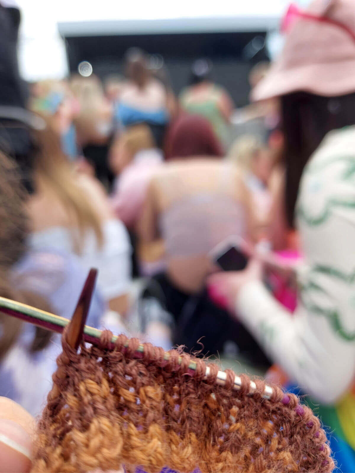 A close up of a partly-knitted sock cuff on a circular needle. In the background are people waiting for a concert to start and the stage behind them. The yarn is Winwick Mum Autumn Leaves in shades of brown