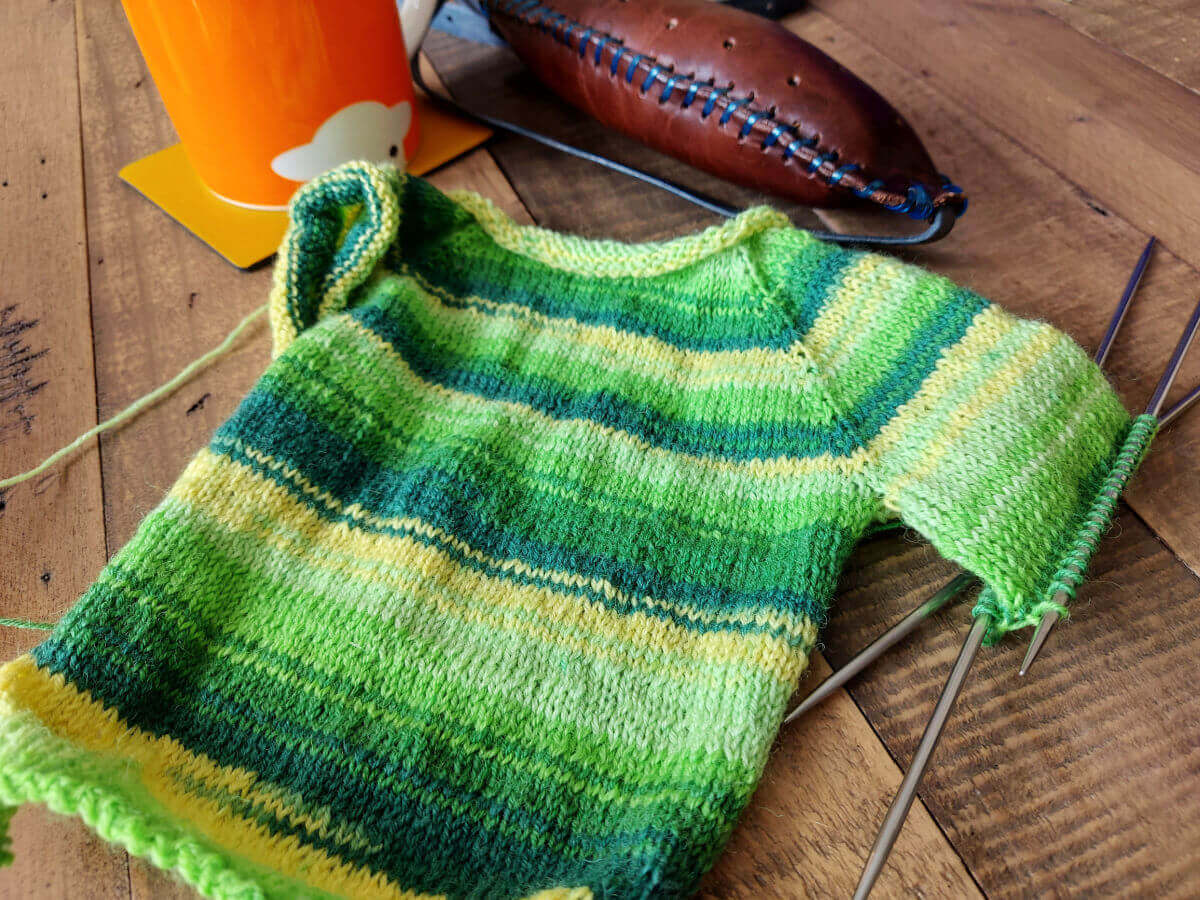 A partly-knitted green and yellow striped baby jacket lying on a wooden table. There is an orange mug and a leather knitting belt next to it