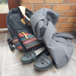 A grey school blazer is lying across a black and colour mosaic-patterned rucksack and a pair of black slip-on shoes. They are all left on a tiled floor against a brick wall.