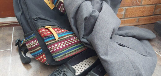 A grey school blazer is lying across a black and colour mosaic-patterned rucksack and a pair of black slip-on shoes. They are all left on a tiled floor against a brick wall.