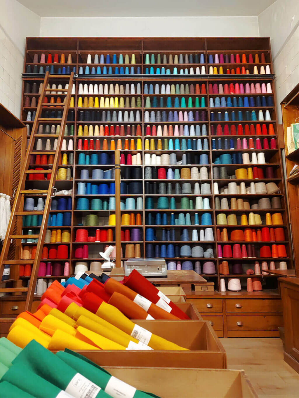 A huge shelving unit containing yarn cones of all colours of the rainbow. There is a ladder to one side of the display to reach the high shelves
