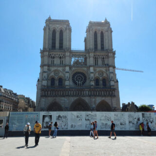 Notre Dame cathedral in Paris. It is being renovated after the fire so is behind hoardings. There is a crane in the background. The bell towers are white against the bright blue sky