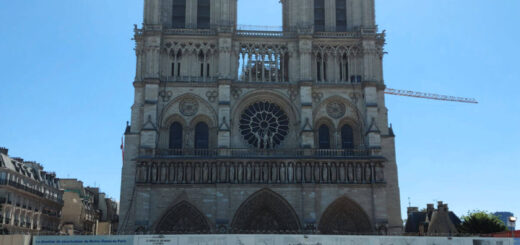 Notre Dame cathedral in Paris. It is being renovated after the fire so is behind hoardings. There is a crane in the background. The bell towers are white against the bright blue sky