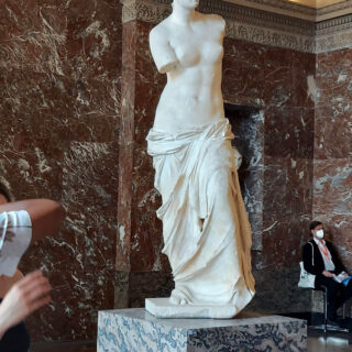 A white marble statue on a mottled marble stand. The statue is of a female, possible Aphrodite, and has no arms.