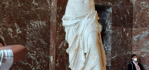 A white marble statue on a mottled marble stand. The statue is of a female, possible Aphrodite, and has no arms.