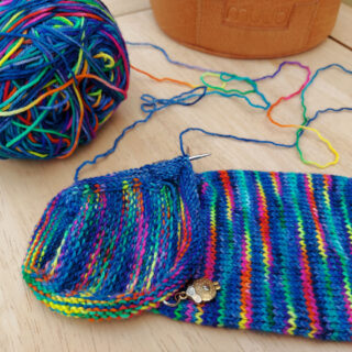 A sock in rainbow colours on a dark blue background is lying on a pale wood table next to the cake of yarn. There are two working yarns on this sock. In the background is a leather project back with the brand name MUUD on it.