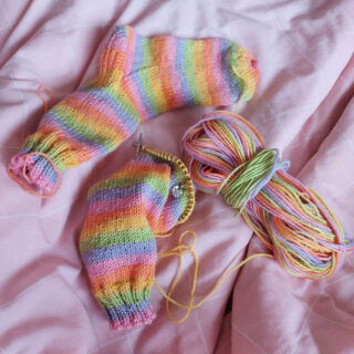 A knitted sock in pale pastel stripes lies next to the half-knitted second sock and the ball of yarn on a pink checked duvet cover