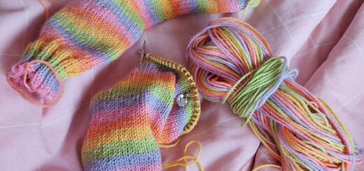 A knitted sock in pale pastel stripes lies next to the half-knitted second sock and the ball of yarn on a pink checked duvet cover