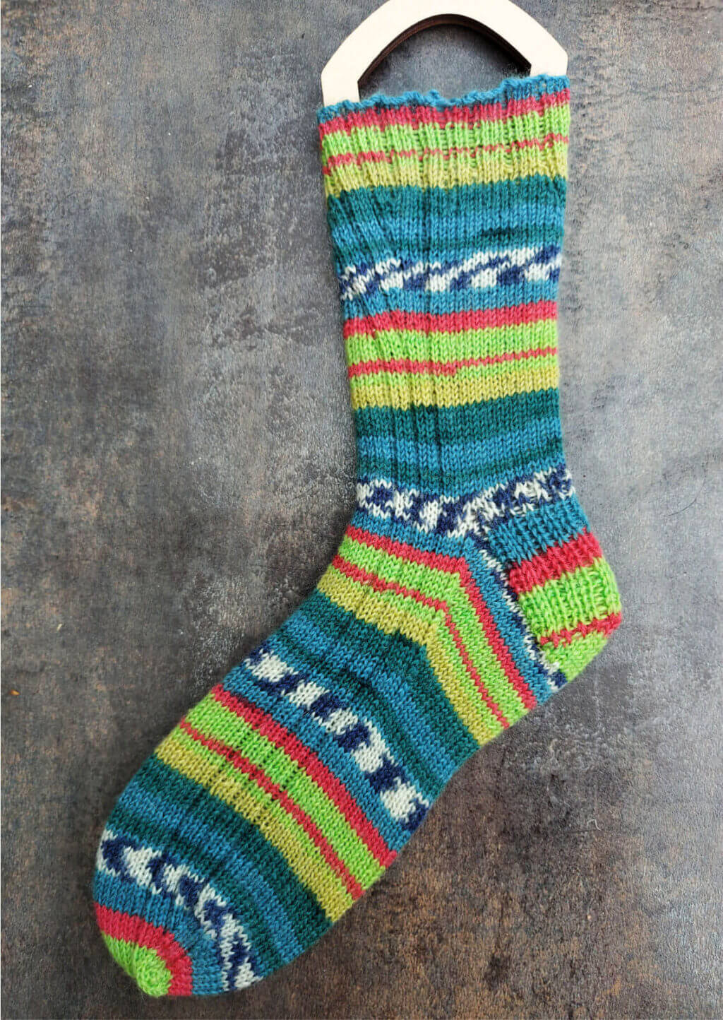 A striped sock on a wooden sock blocker in shades of green, blue and red lies on a brown mottled tile