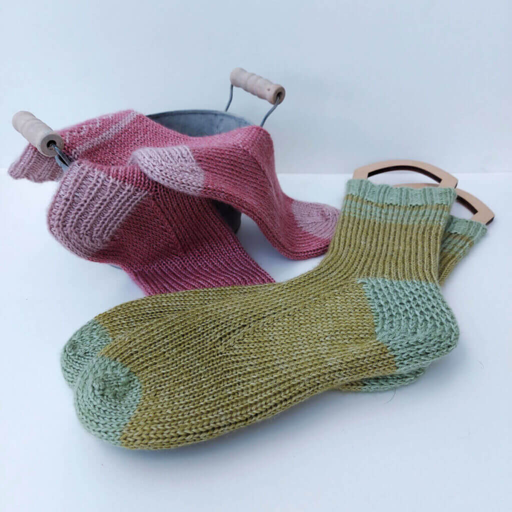 Two pairs of socks - one pink and one green - lie on a cream background. The pink socks are draped over of a decorative metal bucket and the green socks are on wooden sock blockers