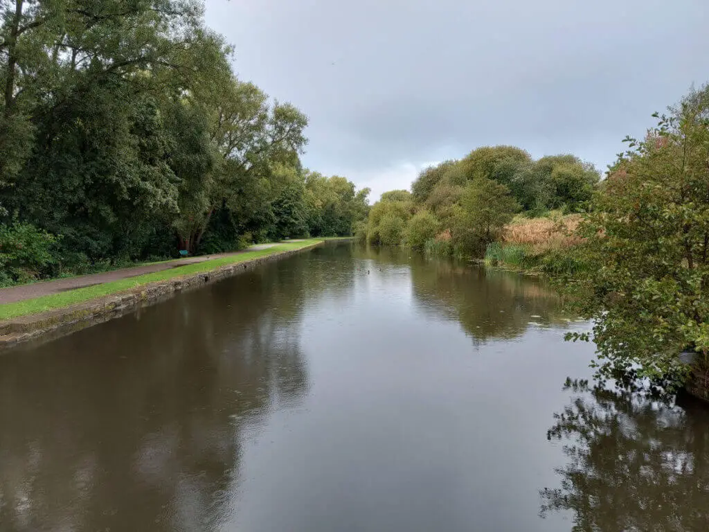 Looking out across a canal from a bridge. The water is very still, there are trees on both sides and grey clouds above. It's raining and you can see it splashing in the water