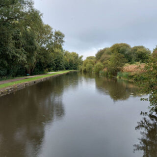 Looking out across a canal from a bridge. The water is very still, there are trees on both sides and grey clouds above. It's raining and you can see it splashing in the water