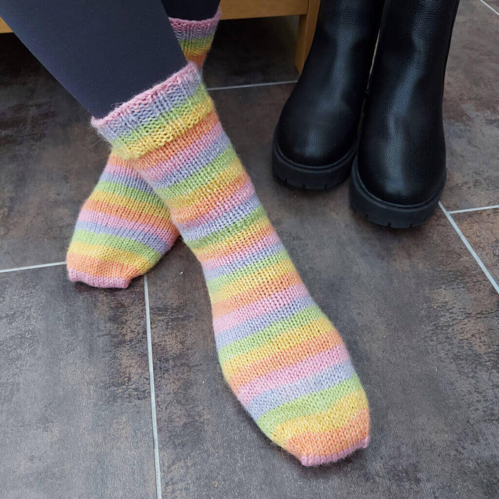 A pair of pale pastel striped socks modelled on feet. The model is sitting on a low wooden table, the floor is tiled in brown tiles and there is a pair of black boots in the top right hand corner of the photo