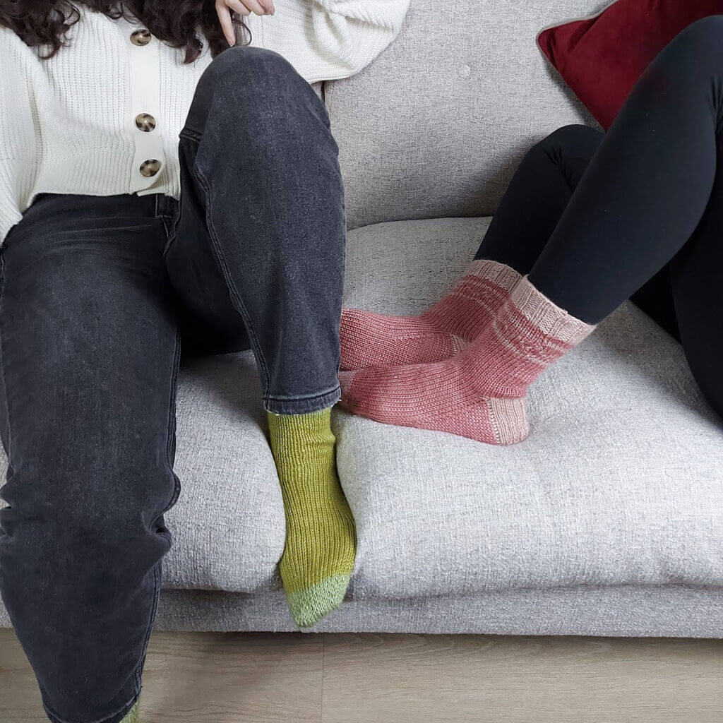 Two young women are sitting on a grey sofa. One is wearing black jeans, green socks and a white cardigan, the other is wearing black leggings and pink socks.