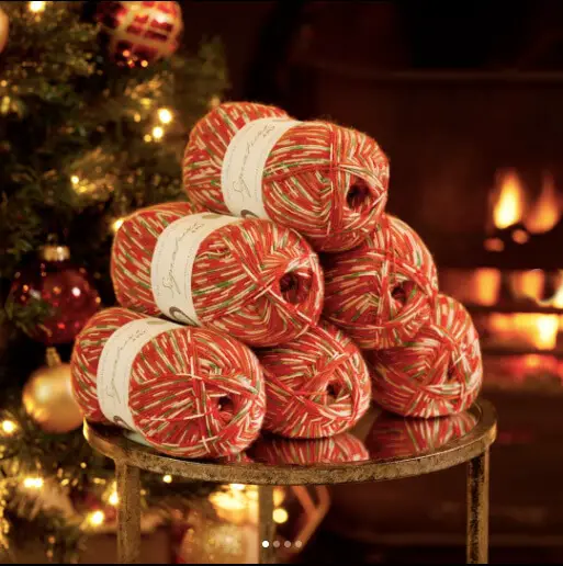 Six balls of WYS Gingerbread yarn are stacked in a pyramid on a small gold-coloured circular table. In the background is a Christmas tree and an open fire