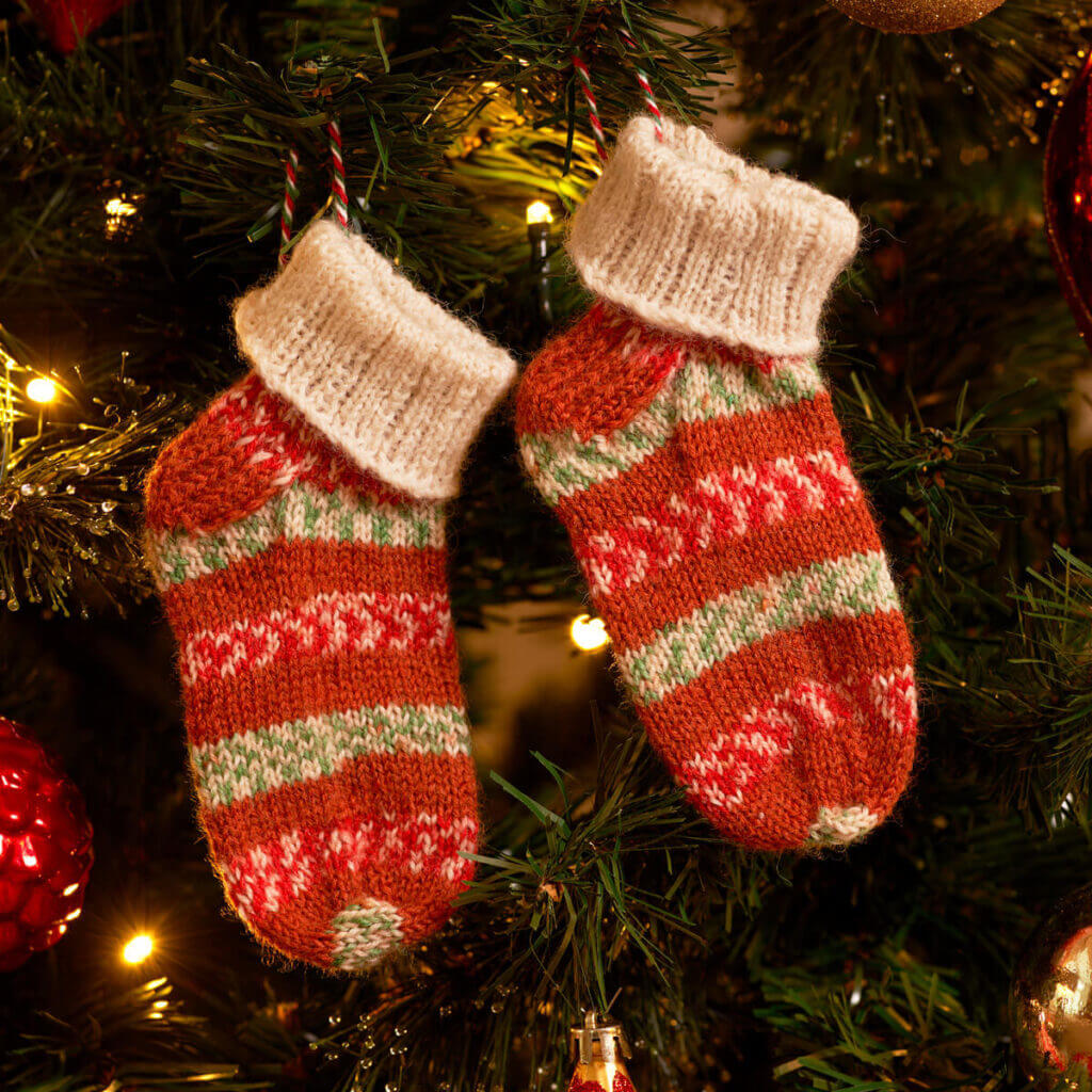 A pair of baby socks in WYS Gingerbread with a rolled cream cuff hang on a Christmas tree