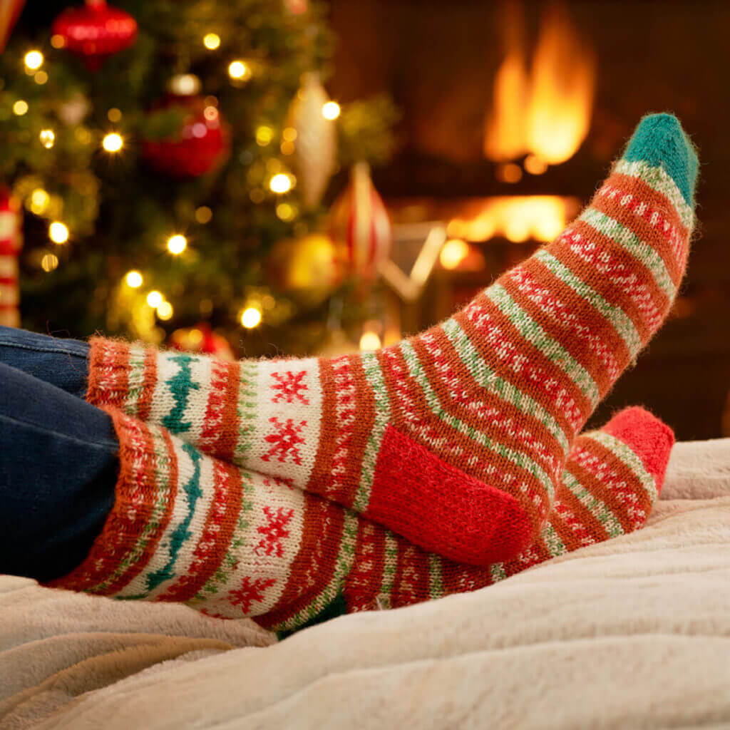 The model is wearing a pair of striped socks with Christmas motifs on the leg. In the background is a Christmas tree and an open fire