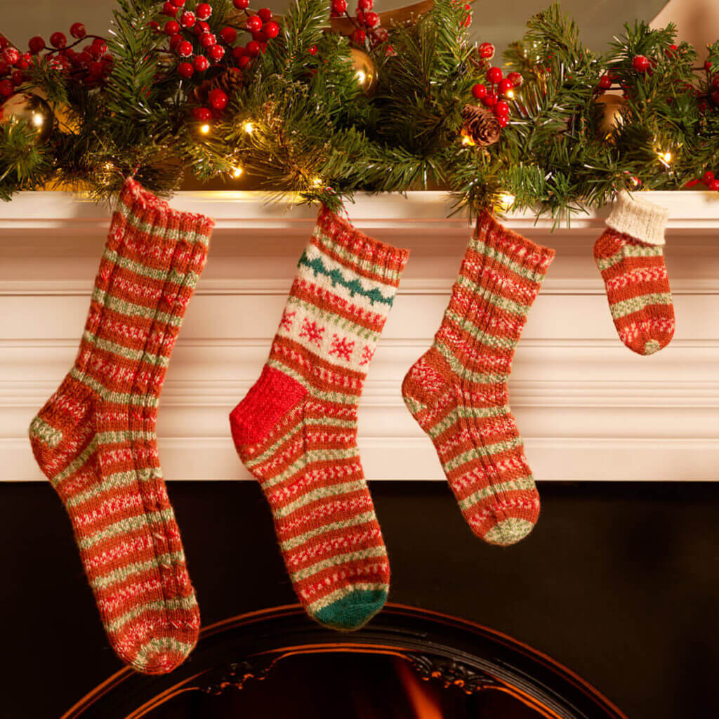 Four knitted socks in sizes from large to baby are hanging on a mantelpiece decorated with Christmas greenery and fairy lights. Each of the socks is a different style but all are knitting in WYS Gingerbread yarn