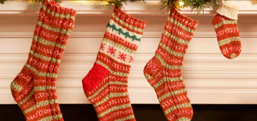 Four knitted socks in sizes from large to baby are hanging on a mantelpiece decorated with Christmas greenery and fairy lights. Each of the socks is a different style but all are knitting in WYS Gingerbread yarn