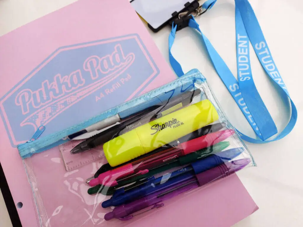 A pad of writing paper with a pink and blue cover lies on a table. On top of the pad is a clear plastic pencil case filled with pens and next to it on the table is a blue lanyard with the word "student" printed on it