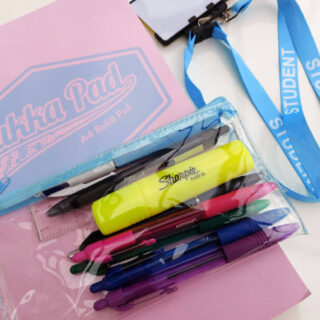 A pad of writing paper with a pink and blue cover lies on a table. On top of the pad is a clear plastic pencil case filled with pens and next to it on the table is a blue lanyard with the word "student" printed on it