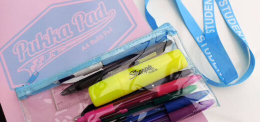A pad of writing paper with a pink and blue cover lies on a table. On top of the pad is a clear plastic pencil case filled with pens and next to it on the table is a blue lanyard with the word "student" printed on it