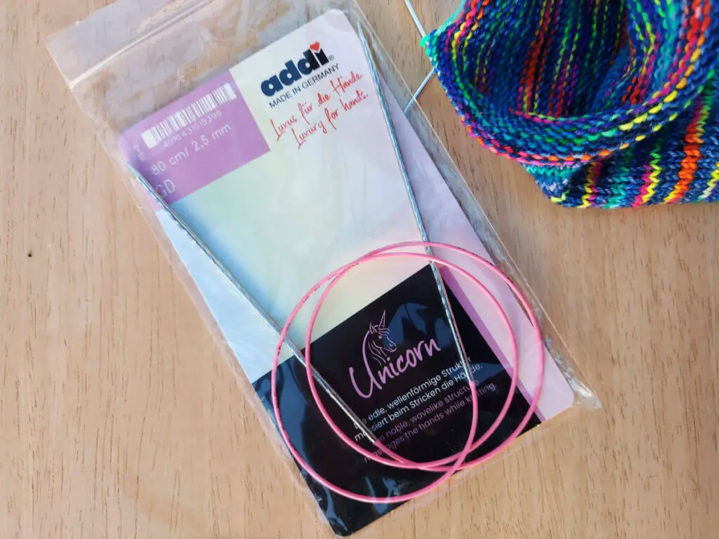 A long circular needle with a pink cable in a packet, lying on a pale wooden table next to a partly-knitted sock