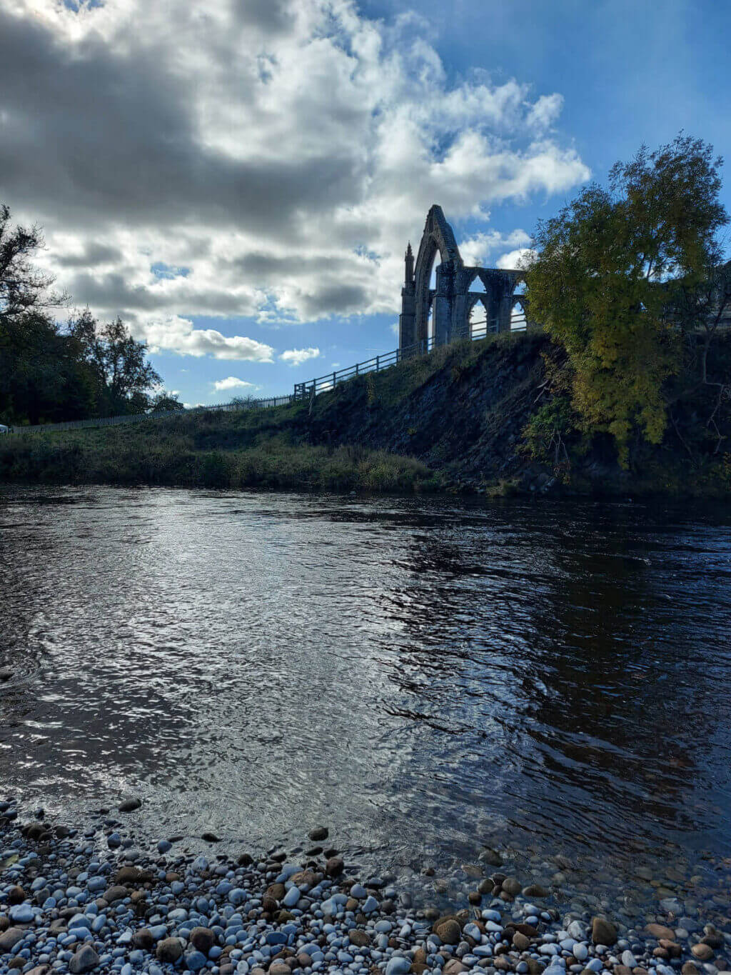 A view across the river to the ruins of Bolton Abbey. It's a bright day but the sun is covered by clouds momentarily and the abbey's stonework looks very dark
